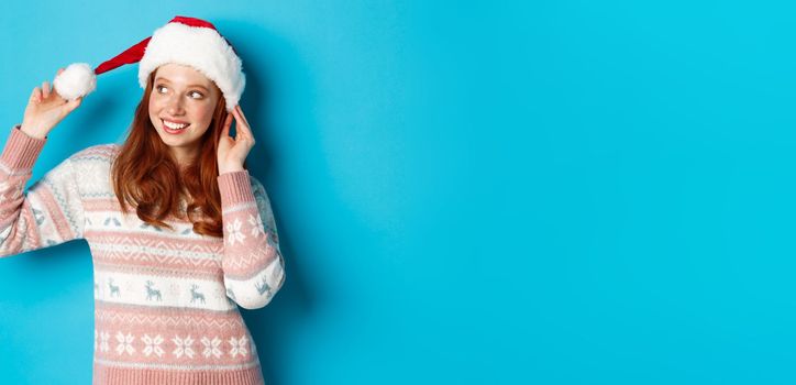 Winter and Christmas Eve concept. Cute teenage girl wearing santa hat and sweater, celebrating xmas, smiling happy, standing over blue background.