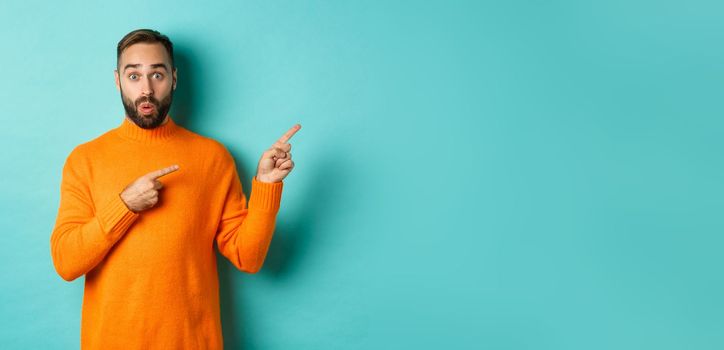 Amazed guy showing advertisement, pointing fingers right at banner, standing against turquoise background.