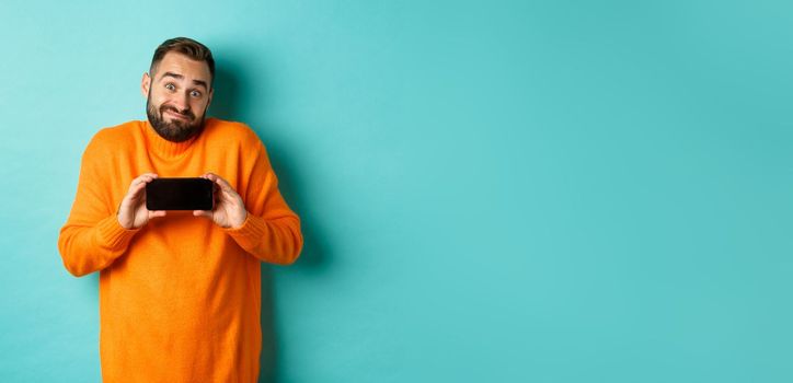 Clueless guy shrugging and showing mobile screen, indecisive emotion, standing in orange sweater over turquoise background.