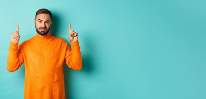 Skeptical and reluctant man looking displeased, pointing fingers up, bad offer, standing over turquoise background.