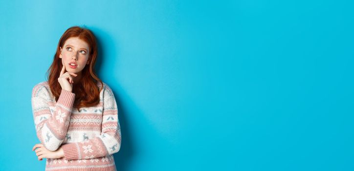 Image of thoughtful redhead girl looking at upper left corner, thinking or making choice, standing in winter sweater over blue background.