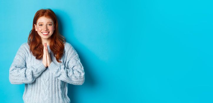 Cute redhead girl saying thank you, smiling and looking at camera, expressing gratitude, standing against blue background.