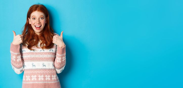 Girl approve something awesome, showing thumb-up, well done gesture and smiling amazed, standing over blue background.