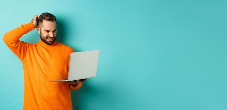 Freelance and technology concept. Confused man scratching head and looking at laptop screen puzzled, dont understand, standing over light blue background.