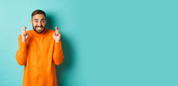 Worried man making a wish, hoping for something, cross fingers good luck, standing nervous over turquoise background.