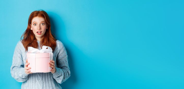 Surprised redhead girl receiving valentines gift, holding box with present and staring at camera amazed, wearing sweater, standing over blue background.