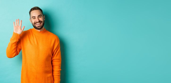 Photo of friendly young man saying hello, smiling and waiving hand, greeting you, standing in orange sweater over light blue background.