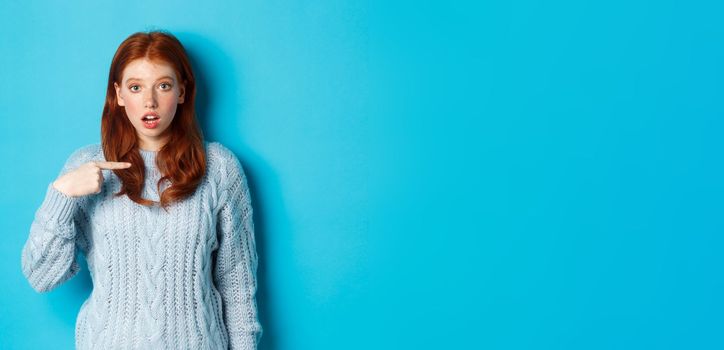 Confused redhead girl pointing at herself, being chosen, standing in sweater against blue background.