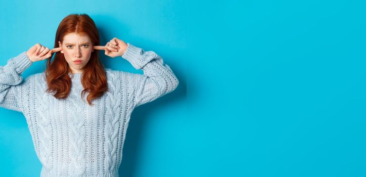 Teenage redhead girl unwilling to listen, shut ears and frowning angry, staring at camera offended, sulking against blue background.