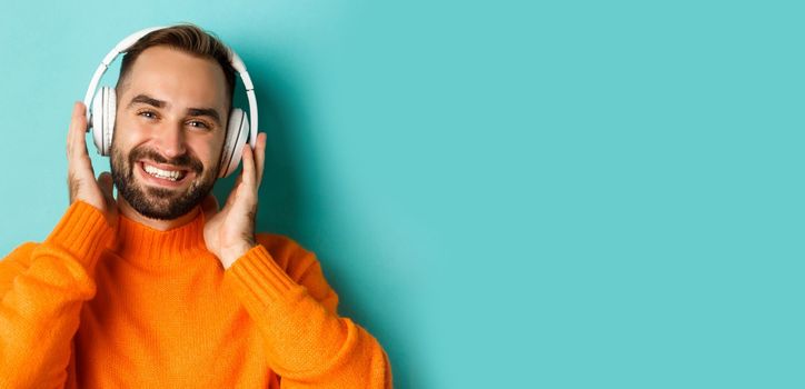 Close-up of handsome modern man listening music in headphones, standing in orange sweater over turquoise background.