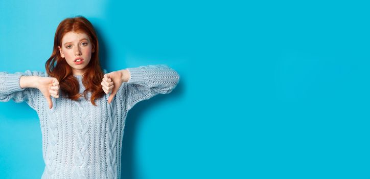 Bored and skeptical redhead girl showing thumbs down, looking unamused and uninterested, standing over blue background.