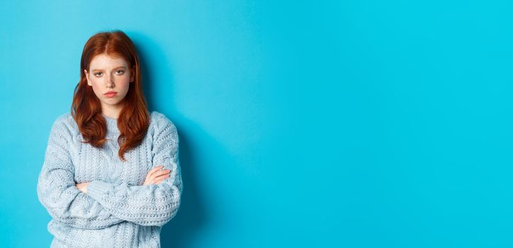 Annoyed and bothered redhead teeange girl cross arms on chest, staring at something lame and boring, standing against blue background.