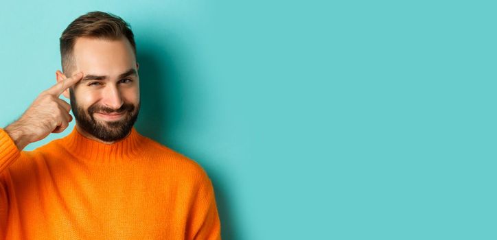 Image of young man in orange sweater, smiling and pointing at head, having an idea, praising good thought, standing over light blue background.