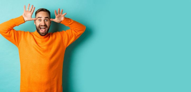 Image of handsome caucasian man making funny faces, mocking someone and smiling, standing against light blue background.