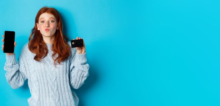 Excited redhead girl showing mobile phone screen and credit card, demonstrating online store or application, standing over blue background.