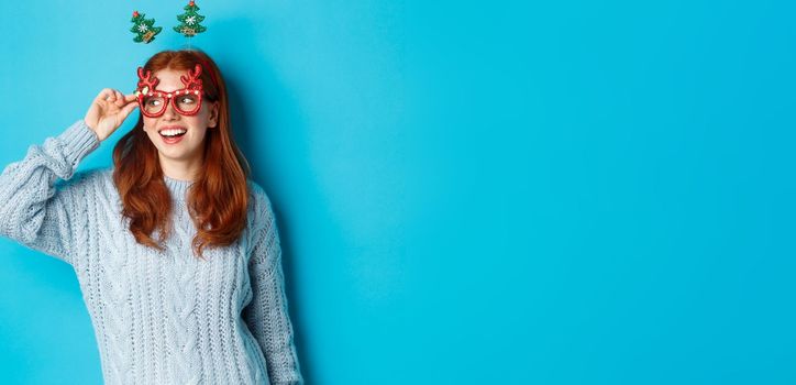 Christmas party and celebration concept. Cute redhead teen girl celebrating New Year, wearing xmas tree headband and funny glasses, looking left amused, blue background.