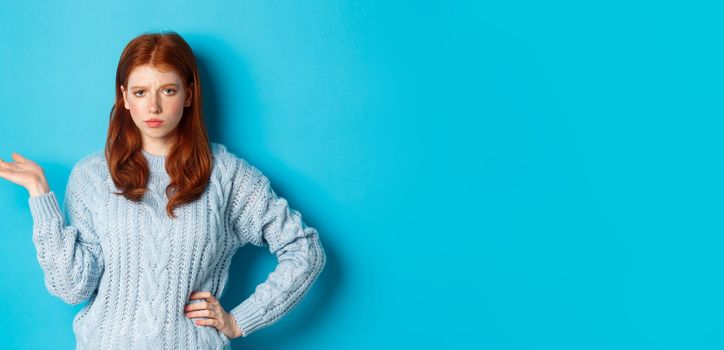 Skeptical teenage girl looking unamused, raising hand in so what gesture, staring at something with careless face, standing over blue background.