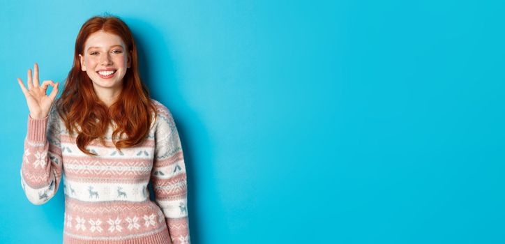 Cute smiling redhead woman showing okay sign, praising good work, well done gesture, standing over blue background.