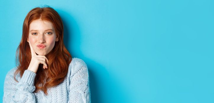 Close-up of beautiful redhead girl thinking, pucker lips and staring thoughtfula t camera, making choice, standing over blue background.