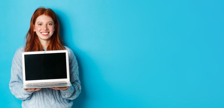 Young smiling woman with red hair and freckles showing computer screen, holding laptop and demonstrate online promo, standing over blue background.