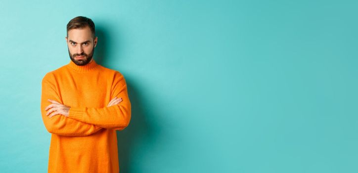 Offended man looking angry at you, cross arms on chest and stare mad, standing in orange sweater against turquoise background.