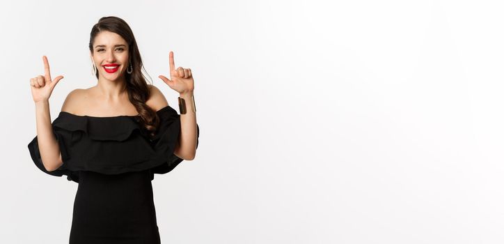 Fashion and beauty. Charming woman with red lips, black dress, smiling happy and pointing fingers up, showing logo, white background.