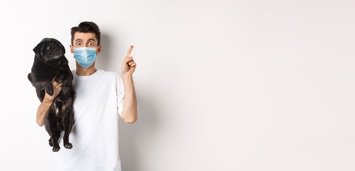 Covid-19, animals and quarantine concept. Young man in medical mask holding cute black pug, dog looking right and owner pointing at logo, white background.