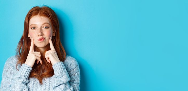 Close-up of cute redhead girl in sweater, pucker lips and pointing fingers at cheeks, poking dimples, standing over blue background.