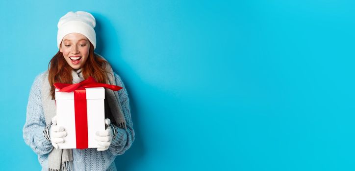 Winter holidays and Christmas eve concept. Surprised cute redhead girl in beanie and sweater receiving New Year gift, looking at present amazed, standing over blue background.