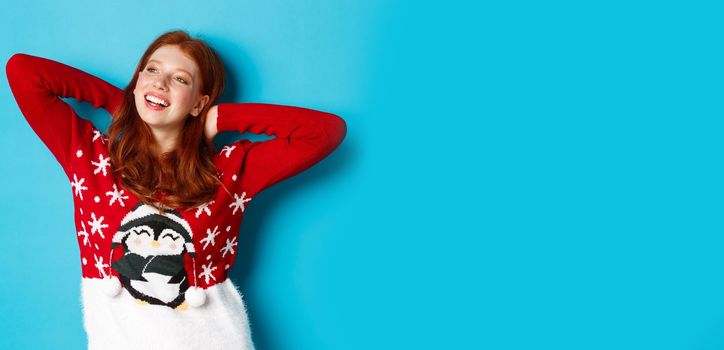 Winter holidays and celebration concept. Relaxed teenage girl holding hands behind head and looking left at promo, resting on Christmas Eve, standing over blue background.