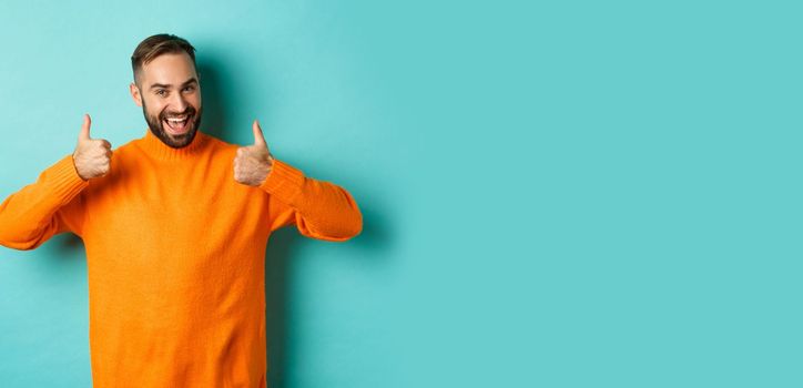 Handsome man expressing support, showing thumb up, encourage you, praise excellent work, approve and agree, standing over light blue background.