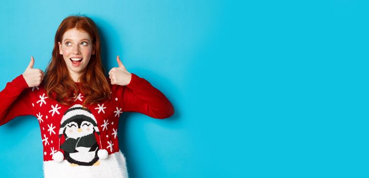 Winter holidays and Christmas Eve concept. Beautiful redhead girl in xmas sweater, celebrating New Year, showing thumbs up and looking at upper left corner, blue background.