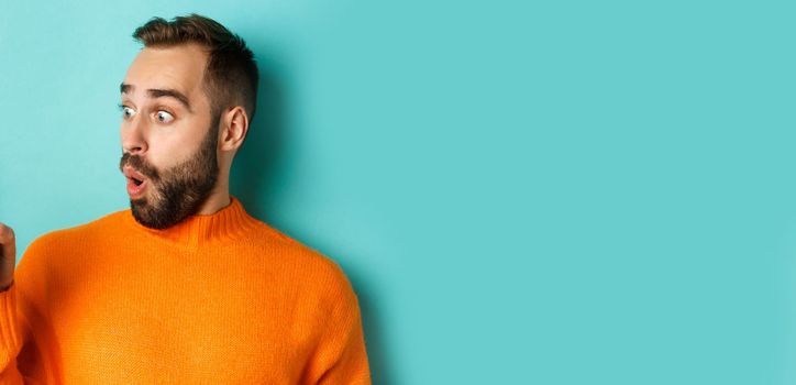 Close-up of young bearded man showing phone screen and looking amazed, wearing orange sweater, standing against studio background.