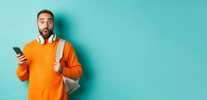 Caucasian man with headphones and backpack staring at camera surprised after reading phone message, standing over turquoise background.