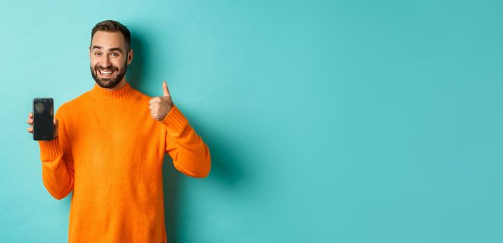 Attractive bearded man showing smartphone screen, thumbs up, recommending mobile app, standing satisfied over turquoise background.