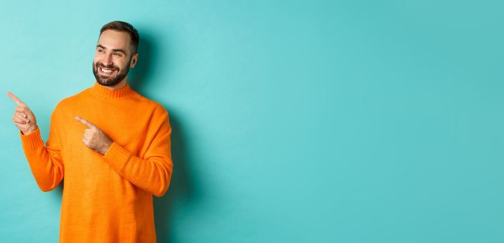 Attractive bearded male model in orange sweater, smiling satisfied and pointing fingers left, standing over turquoise background.