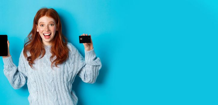 Excited redhead girl showing mobile phone screen and credit card, demonstrating online store or application, standing over blue background.