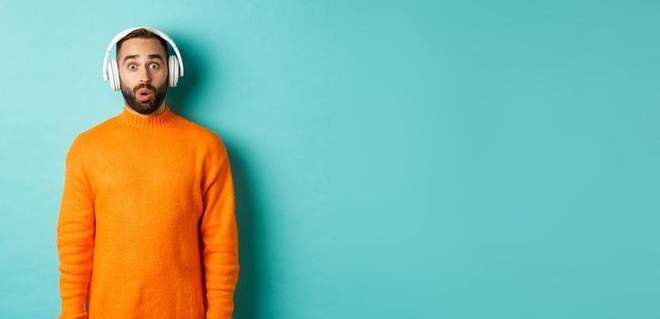 Amazed adult man listening music in headphones, looking at camera impressed with sound, standing over turquoise background.