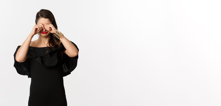 Fashion and beauty. Sad and gloomy woman in black dress crying, rubbing eyes and sulking, standing over white background.