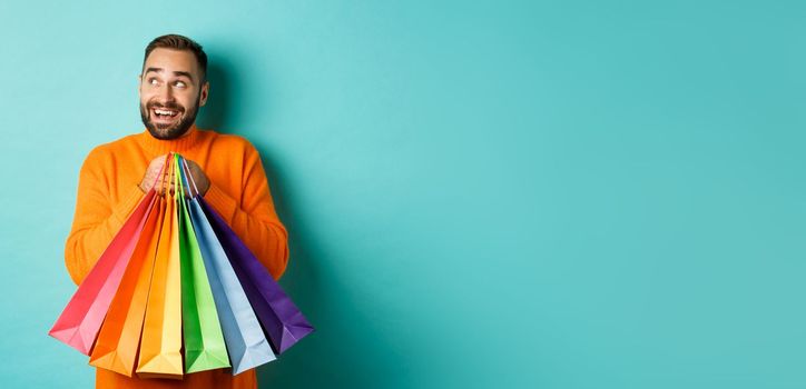 Happy caucasian man in orange sweater, looking left and imaging, holding shopping bags and smiling, standing over turquoise background.