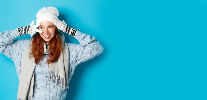 Winter and holidays concept. Cute redhead teen girl in beania, gloves and sweater showing peace sign, looking left at copy space on blue background.