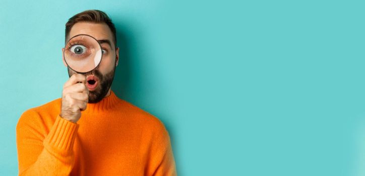 Funny man looking through magnifying glass, searching or investigating something, standing in orange sweater against turquoise background.