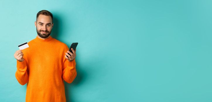 Online shopping. Handsome caucasian man in orange sweater, using credit card and mobile phone, standing over light blue background.