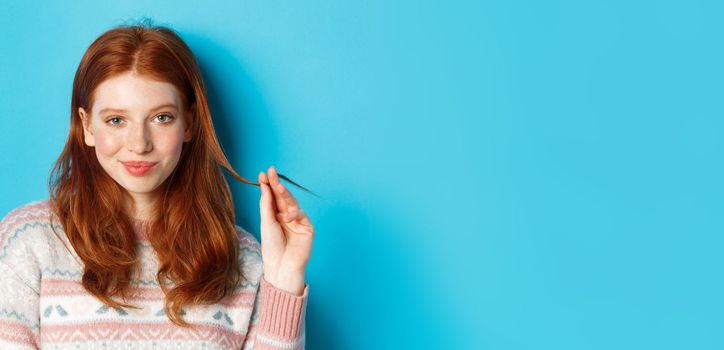 Close-up of confident and sassy redhead teen girl looking at camera pleased, playing with hair strand and smirking, standing over blue background.