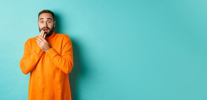 Scared and shocked guy looking at camera, standing in orange sweater against turquoise background.