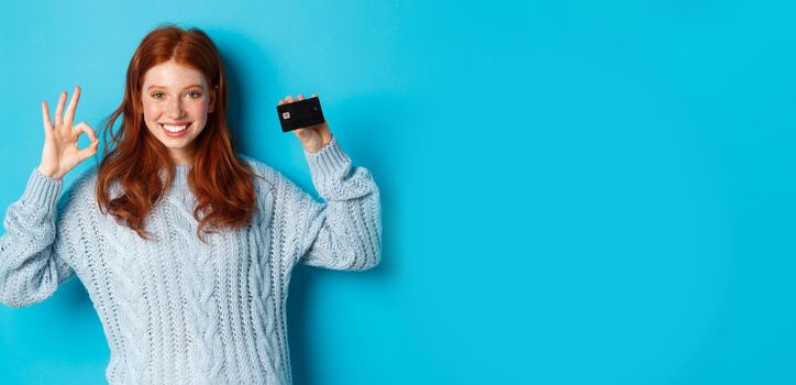 Happy redhead girl in sweater showing credit card and okay sign, recommending bank offer, standing over blue background.