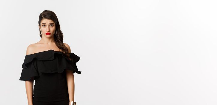 Fashion and beauty. Cute and timid young woman in black dress looking confused and sad at camera, cant understand, standing gloomy against white background.