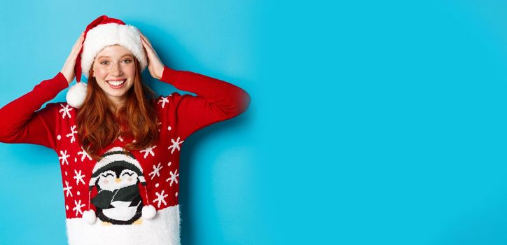 Happy holidays and Christmas concept. Pretty redhead girl in xmas sweater, put on santa hat and smiling, celebrating New Year, blue background.