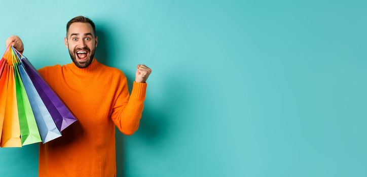 Happy handsome man holding shopping bags and smiling, feeling excitement from discounts, making fist pump, standing over turquoise background.