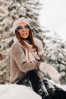 A girl in a sweater and glasses in winter sits on a snow-covered background in the forest.